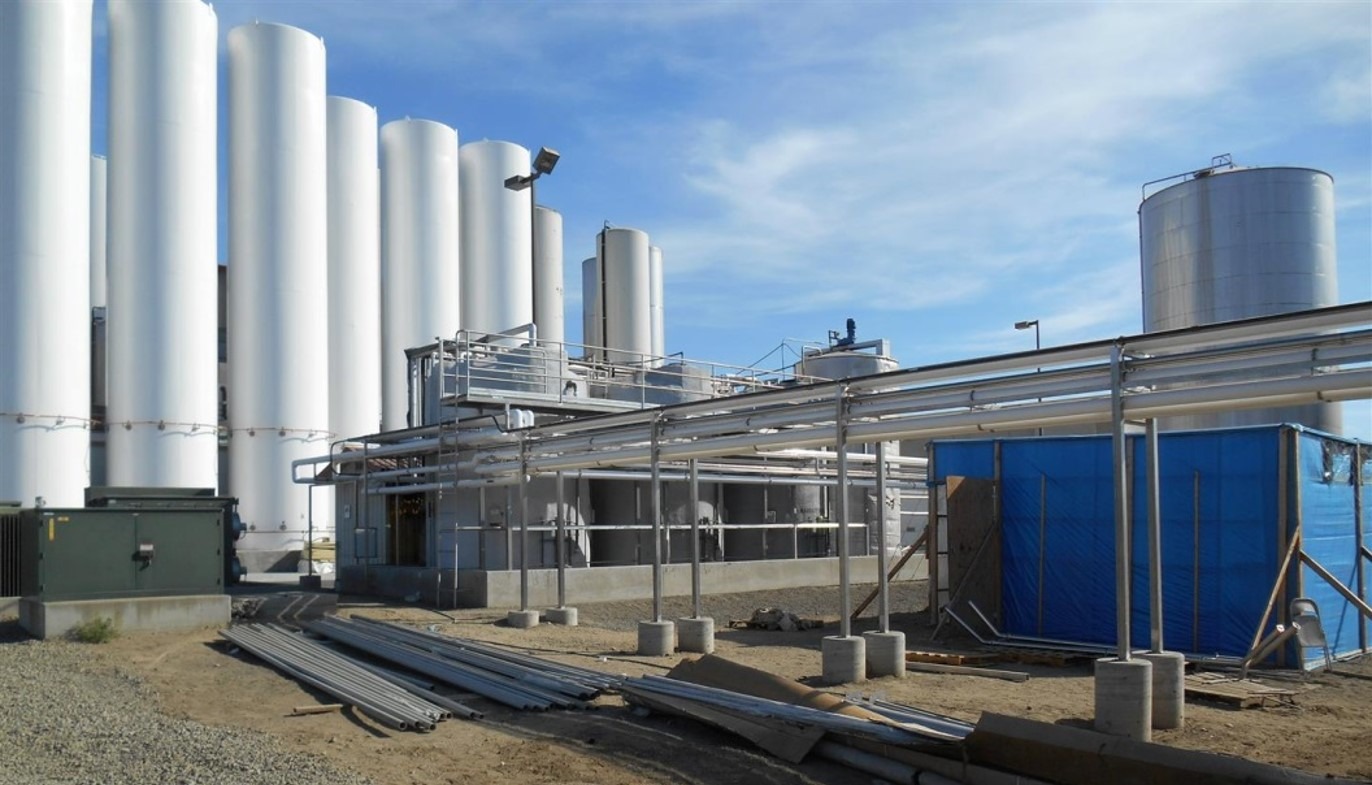 Industrial site featuring tall, white silos and piping system, with a construction area on the right, indicating ongoing operations or upgrades.