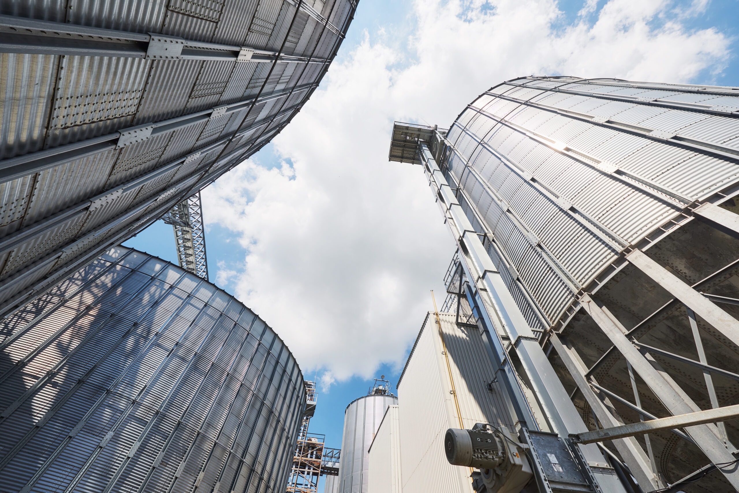 Vertical view of large metal silos against a blue sky with clouds, illustrating industrial grain storage and processing facilities.