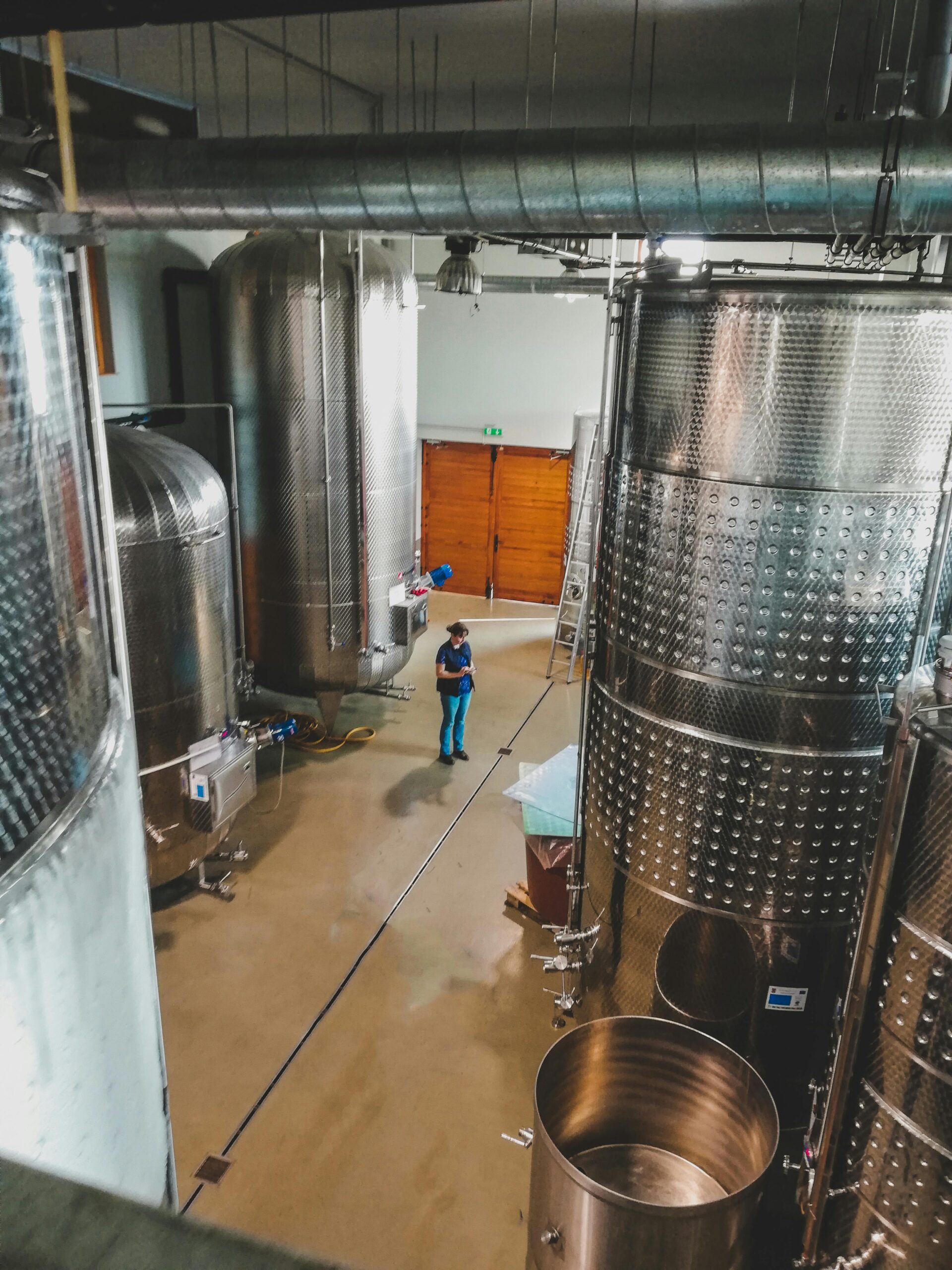 A worker in a blue uniform inspects stainless steel fermentation tanks in a modern winery. The setting highlights the winemaking process.