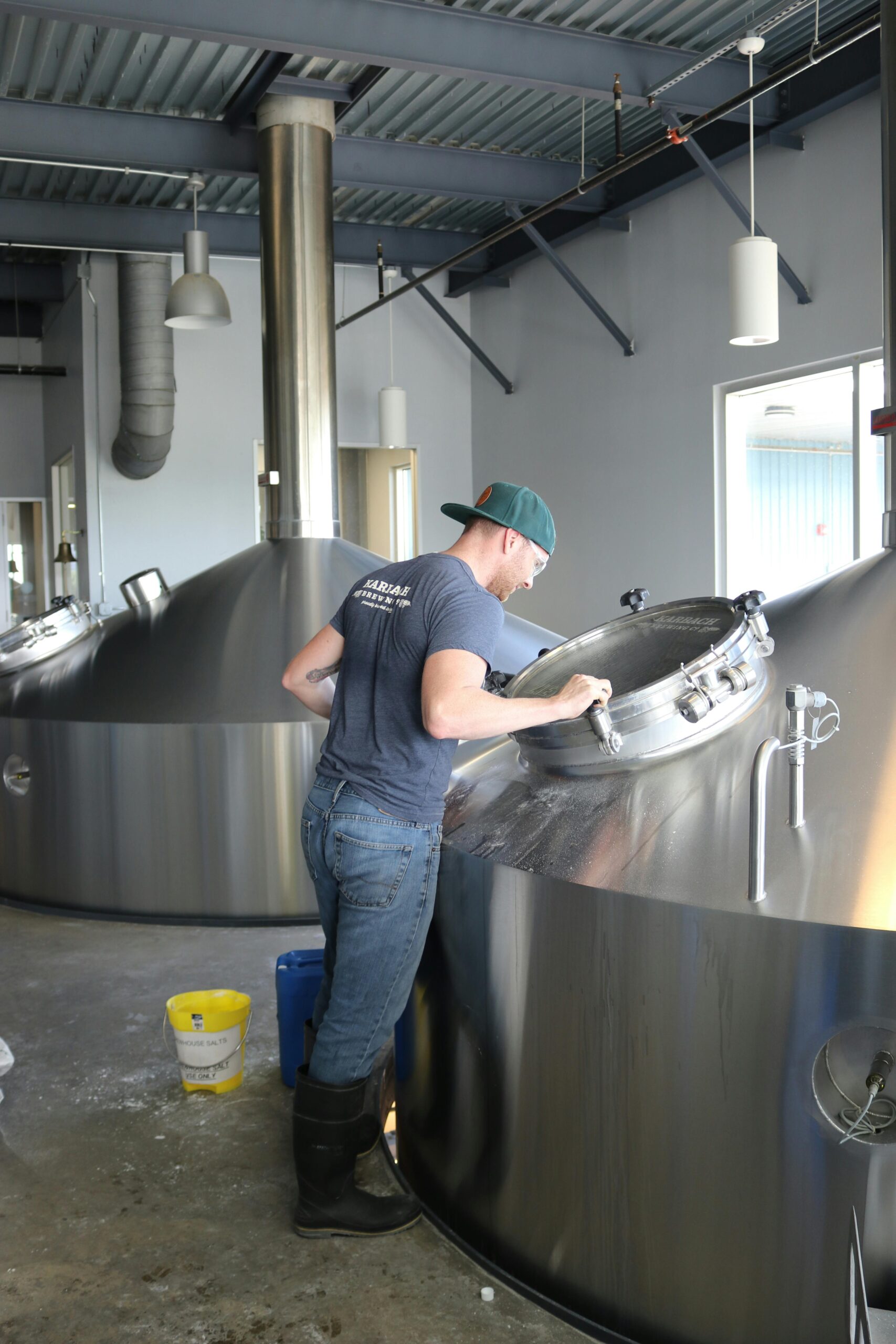 A brewery worker inspects a large stainless steel fermentation tank, surrounded by brewing equipment, emphasizing the production process.
