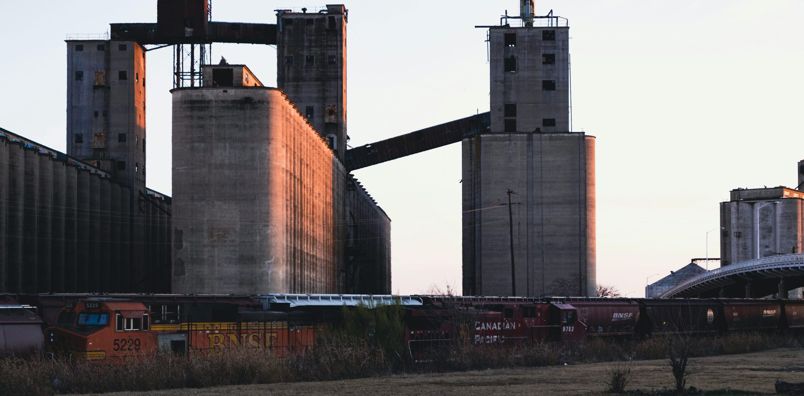 Grain silos and freight trains are silhouetted against a sunset sky, highlighting an industrial landscape crucial for shipping and agriculture.