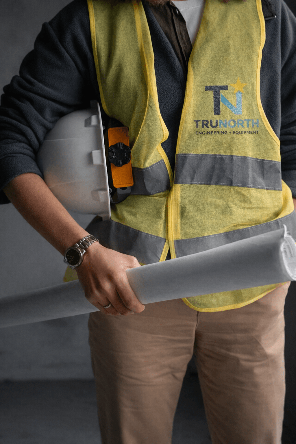 A person in a yellow safety vest holds a white hard hat and rolled blueprints. The vest has the logo "Trunorth Engineering Equipment."