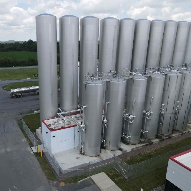 Aerial view of multiple large, silver metal storage tanks arranged in rows next to a small white building with a red trim, surrounded by a fence.